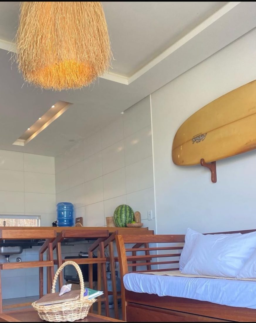 Sala de estar com sofá, prancha de surf na parede e cozinha ao fundo